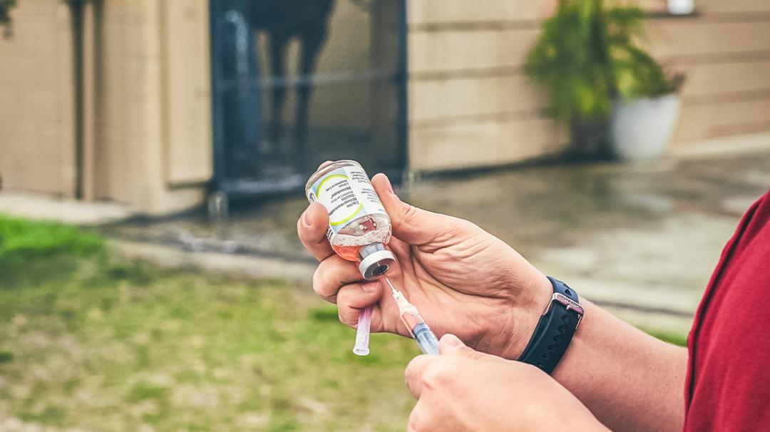 Person drawing liquid into a syringe from a vaccine vial in an outdoor setting.