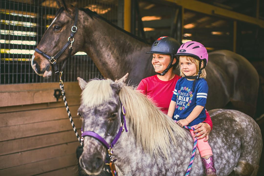 Woman giving education and walking with child riding a horse at a stable.