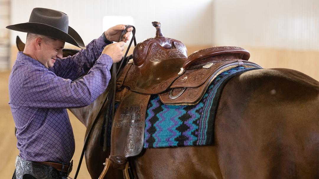 Man in a cowboy hat adjusting the bridle on a horse with a Western saddle in an indoor arena.