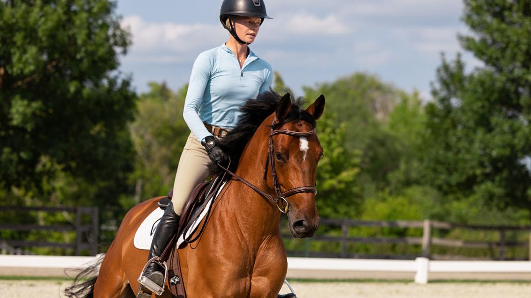 Equestrian riding a bay horse in an outdoor arena, wearing a helmet and light blue shirt.
