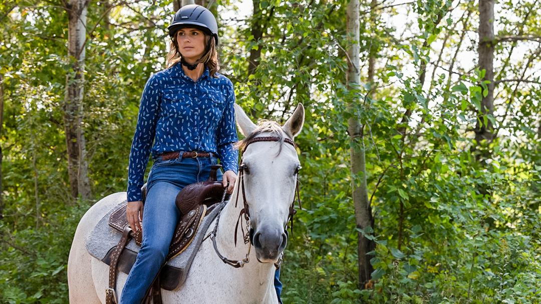 Woman riding a white horse in a Western saddle, surrounded by a forested area.